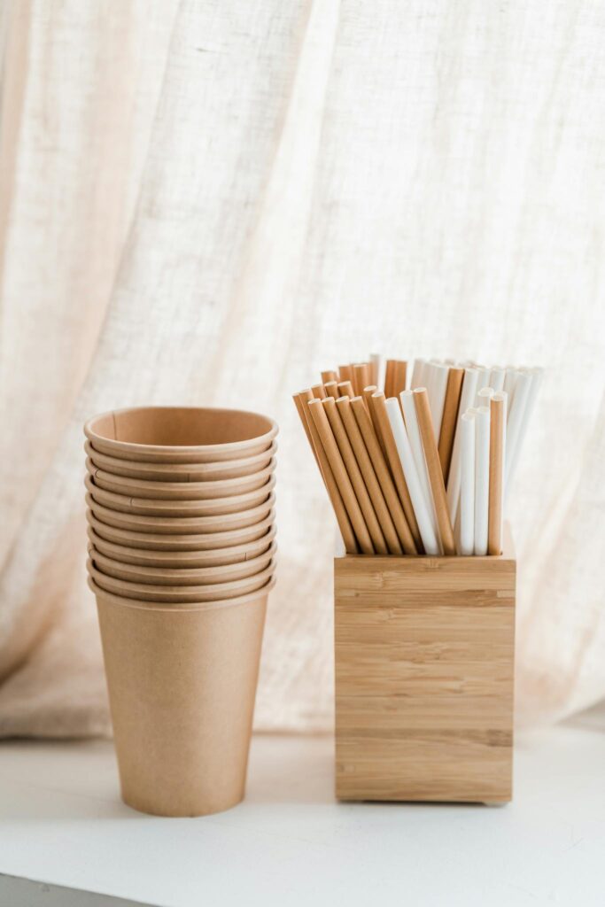 Stacked eco-friendly paper cups and bamboo straws organized in a wooden box against a soft fabric backdrop.