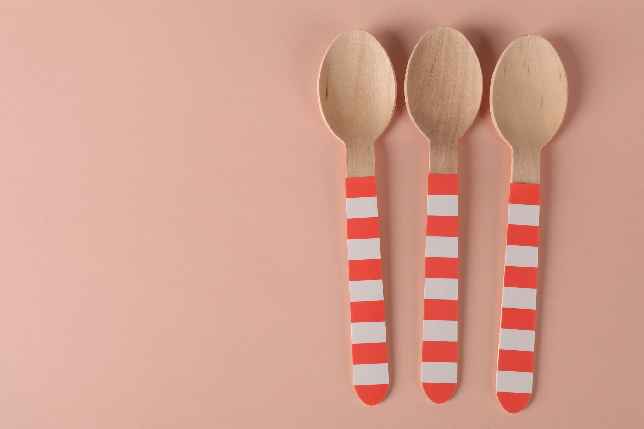 Three eco-friendly wooden spoons with striped handles on a minimalist pink background.